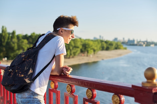 Retrato al aire libre de chico guapo adolescente de 15, 16 años, con espacio de copia. Chico con corte de pelo de moda en gafas de sol con mochila mira el río, de pie en el puente, día soleado de verano al atardecer