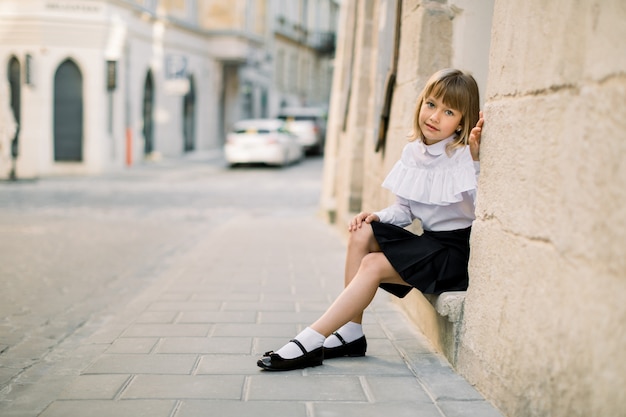 Retrato al aire libre de la ciudad de la calle de la moda de la niña caucásica. Niña feliz sentada cerca de la pared del edificio antiguo en la antigua ciudad europea con puerta azul en el fondo
