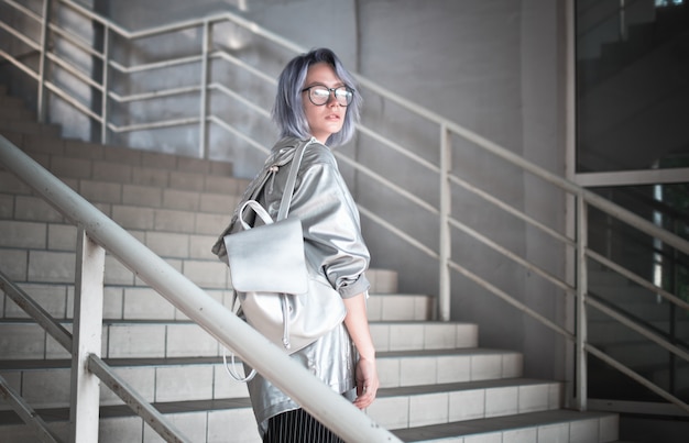 Retrato al aire libre de una estudiante extraordinaria con el pelo azul ceniciento vestido con una capa con una tela plateada y una mochila sobre sus hombros.