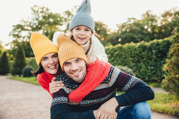 Retrato al aire libre de un hombre guapo que lleva a cuestas a su esposa e hija, usan ropa abrigada, tienen expresiones felices, se apoyan mutuamente, la familia se abraza al aire libre, sonríe agradablemente a la cámara.