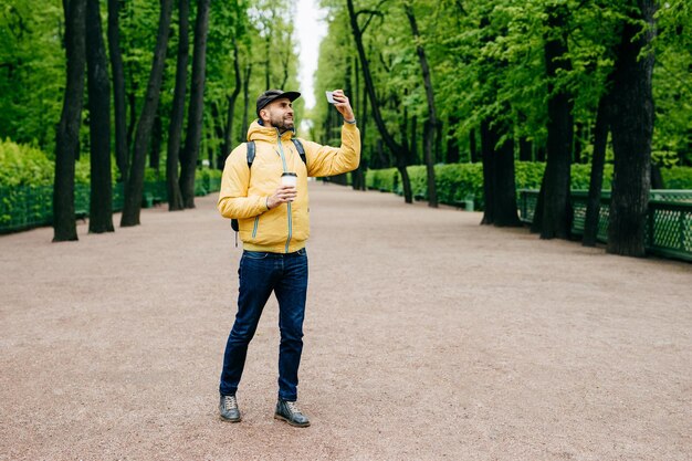 Retrato al aire libre de un joven y apuesto turista vestido con ropa elegante con mochila y café para llevar posando en la cámara de su teléfono inteligente haciendo selfie aislado sobre el fondo verde de la naturaleza