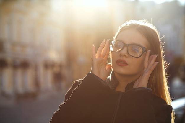 Retrato al aire libre de una maravillosa mujer rubia con gafas y abrigo, posando en la vieja calle soleada. Espacio para texto