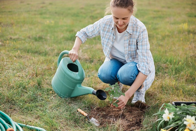 Retrato al aire libre de una mujer con camisa y jeans plantando una joven planta de frambuesa hembra regando el suelo antes de plantar frambuesa al aire libre trabajando en primavera