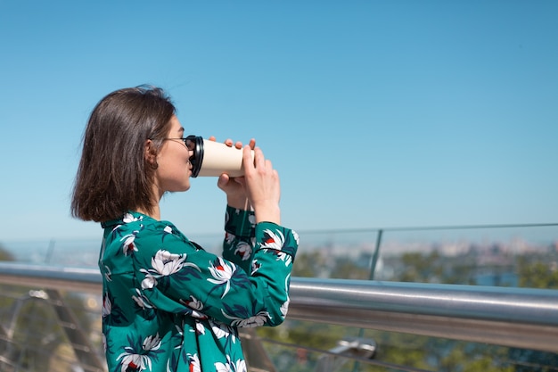 Retrato al aire libre de mujer con camisa verde con una taza de café disfrutando del sol, se encuentra en el puente con una vista increíble de la ciudad en la mañana