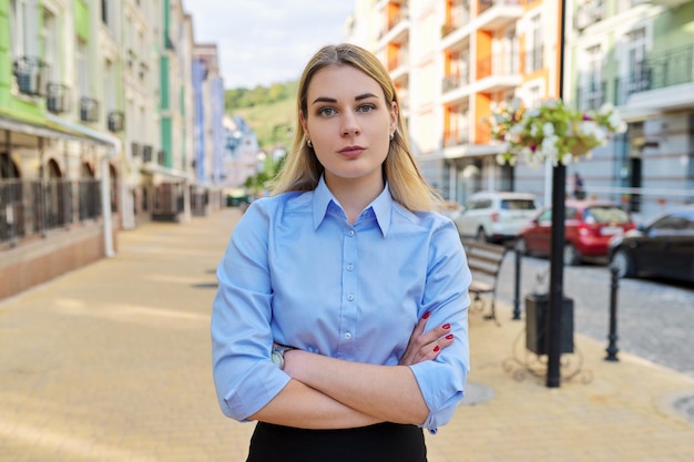 Retrato al aire libre de la mujer de negocios confiada joven con los brazos cruzados, fondo de la calle de la ciudad. Oficinista femenina en camisa azul mira a la cámara