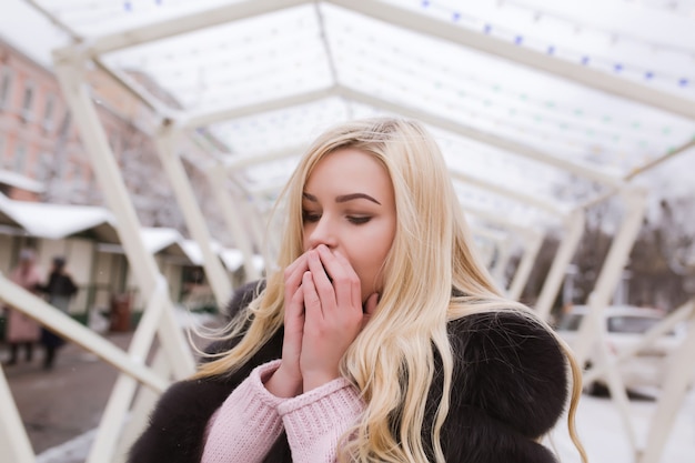 Retrato al aire libre de la mujer rubia joven vestida con estilo calienta sus manos en la calle en invierno