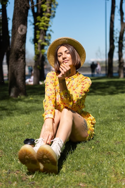 Retrato al aire libre de mujer en vestido amarillo de verano y sombrero sentado sobre el césped en el parque