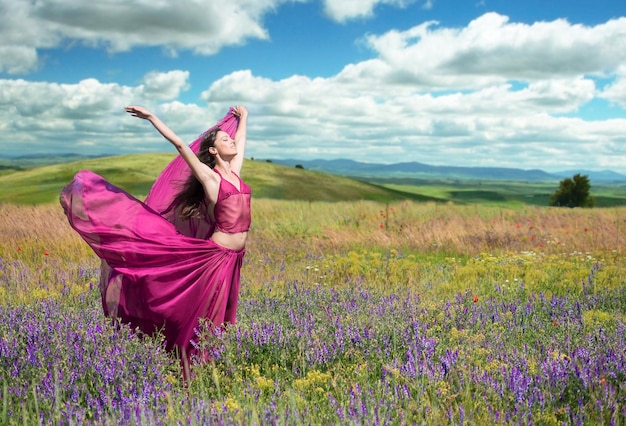 Retrato al aire libre de una mujer con un vestido carmesí aireado posando en el campo floreciente en un día ventoso de primavera