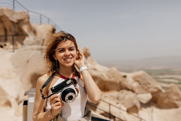 Retrato al aire libre de una niña feliz sonriente sosteniendo una cámara retro y caminando entre rocas antiguas en la luz del sol de cerca