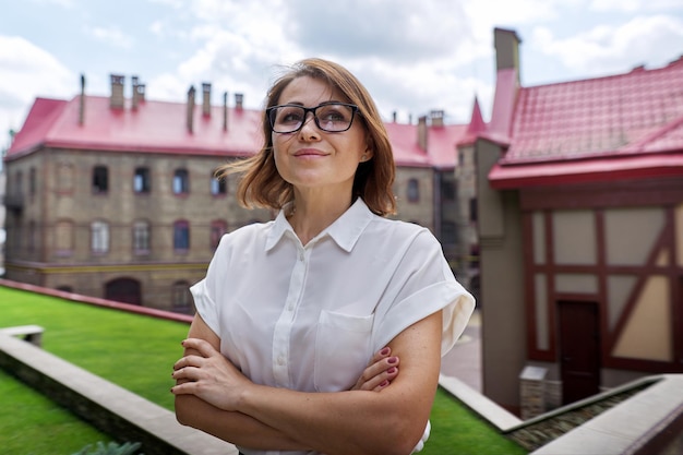 Retrato al aire libre de la sonriente mujer de negocios madura confiada con los brazos cruzados. Exitosa líder femenina de mediana edad en gafas con camisa ligera