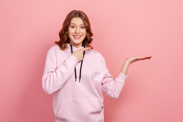 Retrato de una alegre adolescente de pelo rizado con capucha apuntando con el dedo a la derecha en el espacio vacío de la copia con un lugar de sonrisa con dientes para publicidad Foto de estudio interior aislada en fondo rosa