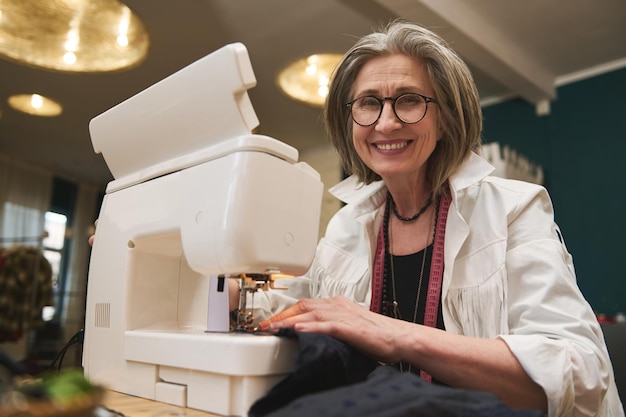 Retrato de una alegre y agradable mujer caucásica de pelo gris diseñadora de moda sastre cose ropa en una máquina de coser en un taller de sastrería