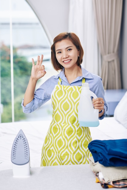 Retrato de alegre ama de casa vietnamita con una botella de agua perfumada para planchar en la mano mostrando signo de ok