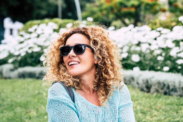 Retrato alegre ao ar livre em uma jovem e feliz mulher bonita sorrindo com flores do parque em segundo plano Atividade de lazer ao ar livre pessoas do sexo feminino desfrutando Usando óculos escuros Ambiente verde