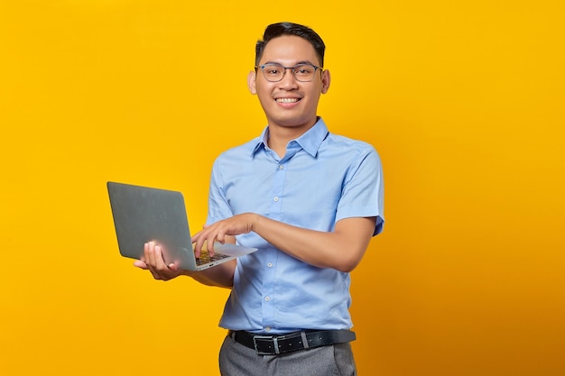 Retrato de un alegre y apuesto hombre asiático con anteojos usando una laptop aislada en un concepto de empresario y emprendedor de fondo amarillo