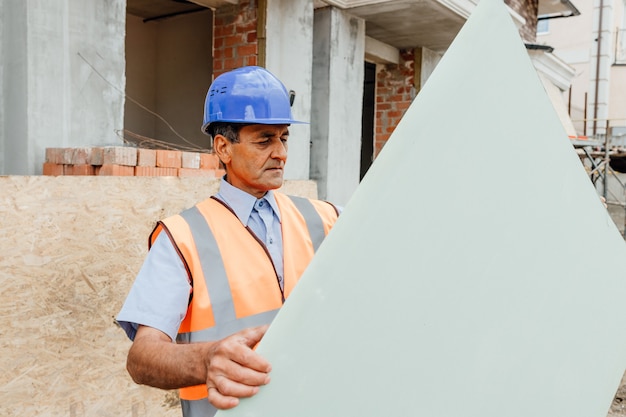 Foto retrato de alegre arquitecto hombre indio americano en el sitio de construcción sostiene un panel de yeso de cartón mirando a la cámara ingeniero civil maduro exitoso en el sitio de construcción con espacio de copia