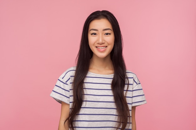 Retrato de una alegre y bonita chica asiática con el pelo largo y moreno en una camiseta a rayas mirando a la cámara con una sonrisa dentuda disfrutando de la vida feliz, un estudio interior de buen humor aislado en un fondo rosa