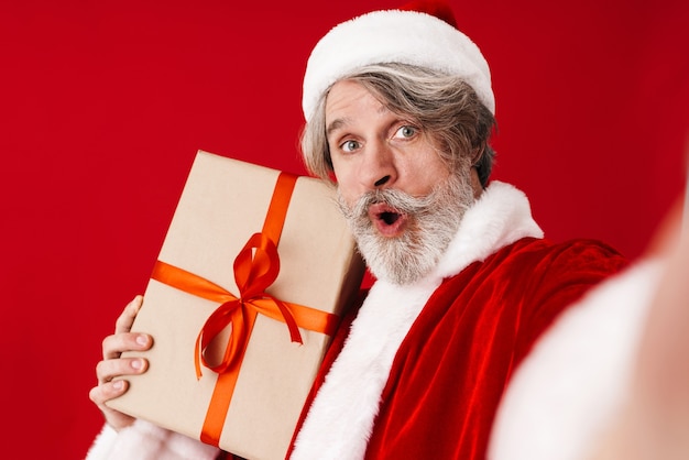 Retrato de alegre canoso viejo hombre de Santa Claus riendo y sosteniendo caja de regalo aislado en rojo en estudio