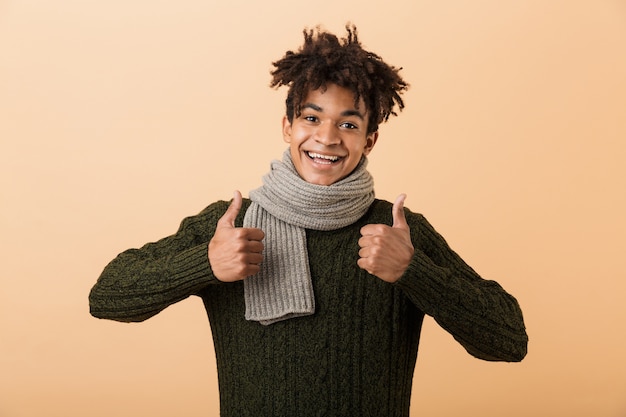 Retrato de alegre chico afroamericano vistiendo suéter y bufanda mostrando los pulgares hacia arriba, aislado sobre pared beige