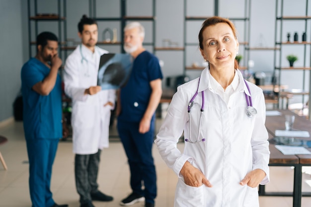 Retrato de una alegre doctora con uniforme blanco parada en la oficina de reuniones médicas, tomándose de la mano en los bolsillos, mirando la cámara. Equipo multirracial de profesionales que trabajan en segundo plano en el hospital.