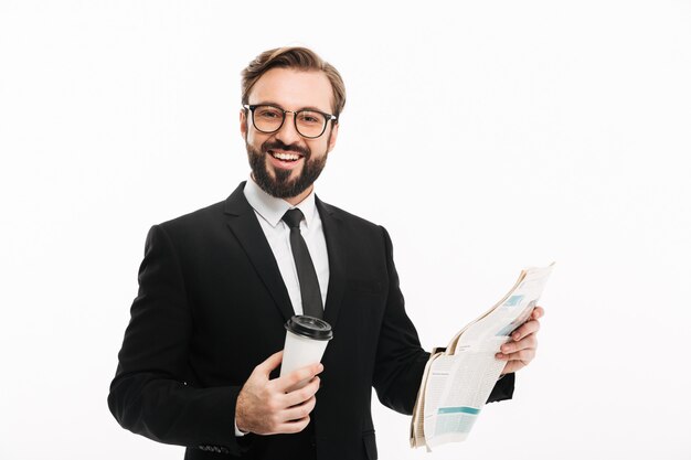 Retrato de alegre empleador masculino en traje y anteojos sonriendo mientras bebe café para llevar y lee el periódico, aislado sobre la pared blanca