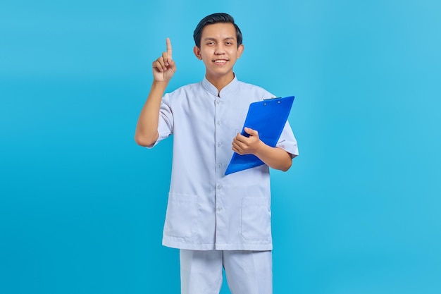 Retrato de alegre enfermero sosteniendo portapapeles de pie y apuntando hacia arriba sobre fondo azul.