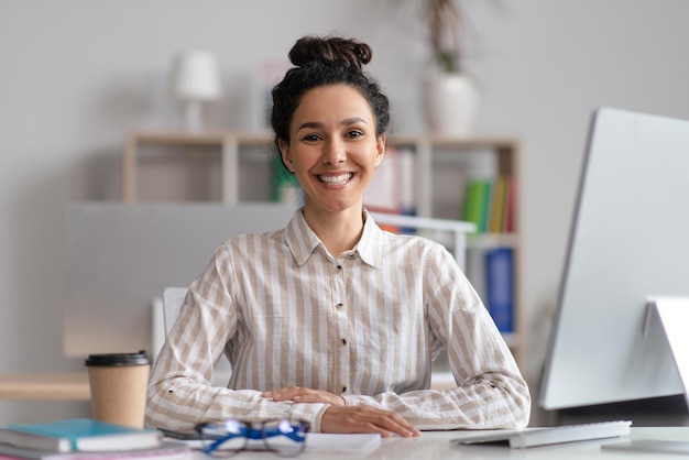 Retrato de una alegre gerente sentada en el lugar de trabajo en una oficina contemporánea y sonriendo a