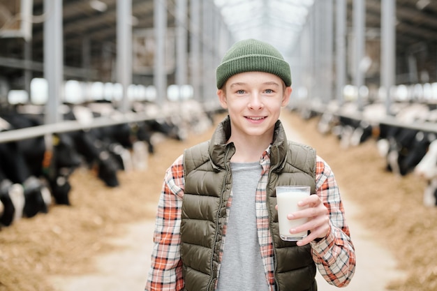 Retrato de alegre granjero con sombrero y chaleco bebiendo leche fresca después de ordeñar la vaca en el establo