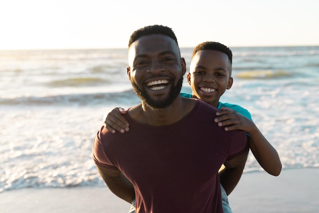 Retrato de un alegre hombre afroamericano llevando a su hijo contra el pintoresco mar y el cielo al atardecer. Copie el espacio, inalterado, familia, juntos, infancia, naturaleza, vacaciones, disfrute y concepto de verano.