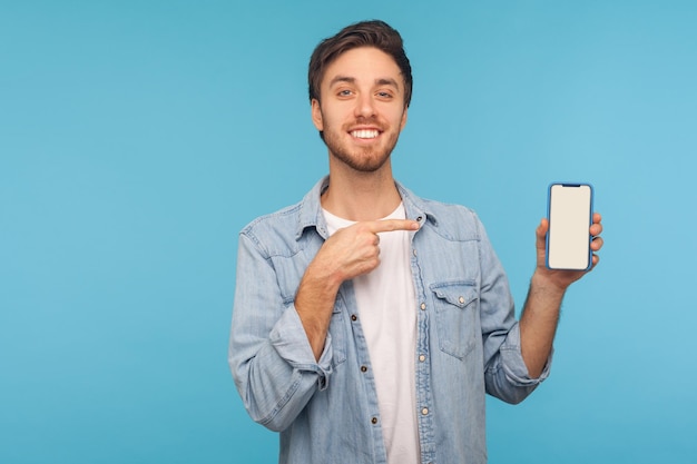 Retrato de un alegre hombre feliz con un pantalones de mezclilla trabajador sonriendo y mostrando un dispositivo móvil, teléfono celular con una pantalla vacía simulada para anunciar. tiro de estudio interior aislado sobre fondo azul, espacio de copia