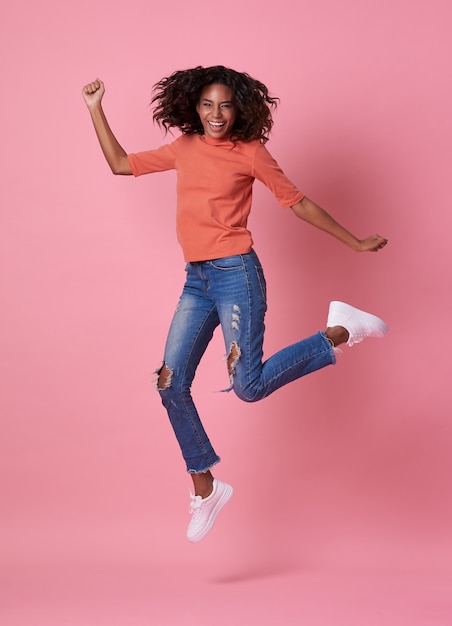 Retrato de una alegre joven africana en camisa naranja saltando y celebrando sobre rosa.
