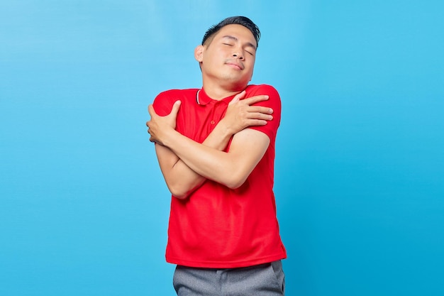 Retrato de un alegre joven asiático con camisa roja abrazándose a sí mismo y sintiéndose frío aislado de fondo azul