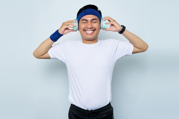 Retrato de alegre joven deportista asiático con diadema azul y camiseta blanca de ropa deportiva mientras escucha música favorita con auriculares aislados en fondo blanco Concepto de deporte de entrenamiento