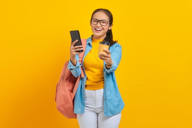 Retrato de una alegre joven estudiante asiática vestida de denim con mochila sosteniendo una taza de café y un smartphone aislado de fondo amarillo