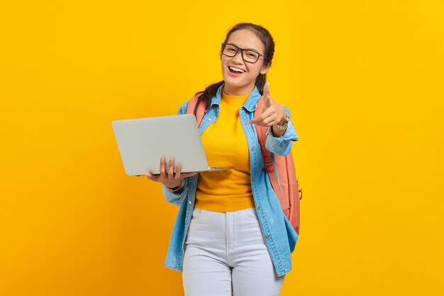 Retrato de una alegre joven estudiante asiática vestida de forma casual con mochila usando una laptop y señalando a la cámara con el dedo aislado en un fondo amarillo Educación en el concepto de universidad universitaria