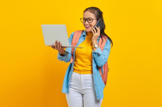 Retrato de una alegre joven estudiante asiática vestida de forma informal con mochila hablando por teléfono móvil y sosteniendo una laptop aislada de fondo amarillo. Educación en concepto de universidad universitaria