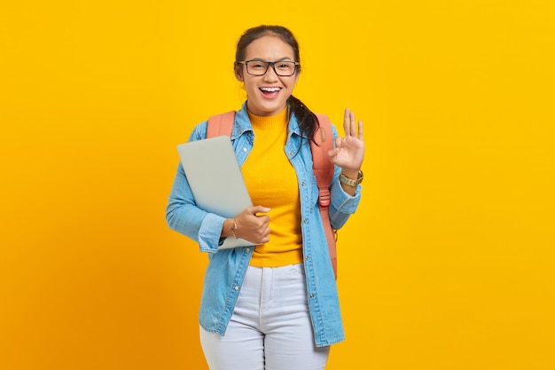 Retrato de una alegre joven estudiante asiática vestida con ropa informal con mochila sosteniendo una laptop y mostrando un gesto correcto con el dedo aislado en un fondo amarillo Educación en el concepto de universidad universitaria