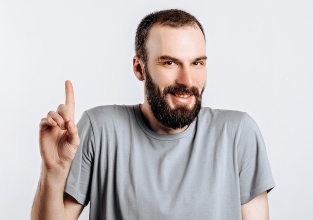 Retrato de alegre joven guapo sonriendo mirando a la cámara apuntando con el dedo hacia arriba sobre la superficie blanca