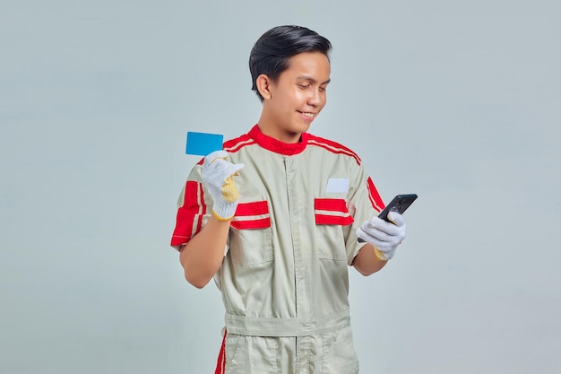 Retrato de alegre joven mecánico mostrando tarjeta de crédito y con smartphone sobre fondo gris