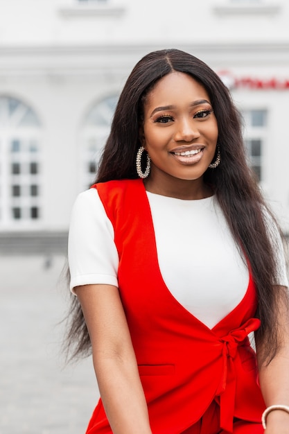 Retrato alegre joven negra con piel limpia y sana con sonrisa encantadora con ojos hermosos en ropa roja de moda al aire libre en la ciudad. Atractiva chica africana sonriente se relaja en la calle.