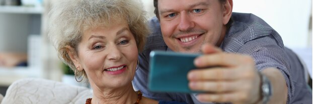 Retrato de alegre madre e hijo tomando selfie para recordar. Sonriente anciana posando para la foto en el sofá en casa. Relación familiar y concepto de tiempo libre.