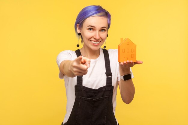 Retrato de una alegre y moderna chica hipster de moda con el pelo corto violeta en overoles de mezclilla sosteniendo una casa de papel naranja y apuntando a la cámara en un estudio interior aislado en un fondo amarillo