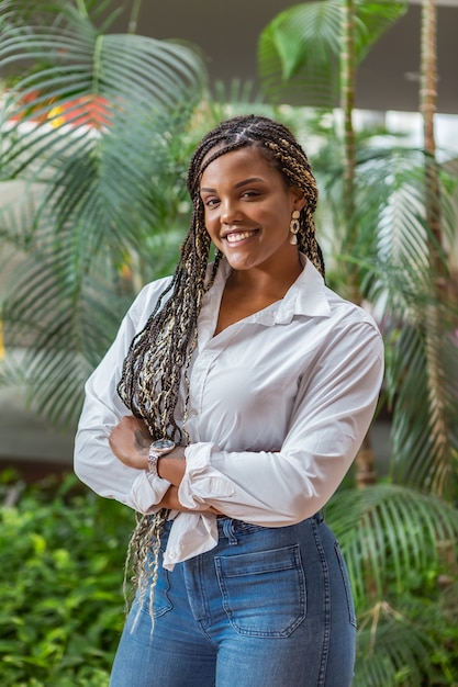 Retrato de una alegre mujer adulta afroamericana de pie con los brazos cruzados y mirando a la cámara. Mujer adulta con trenzas negras y amarillas posando sobre un fondo con plantas y árboles.