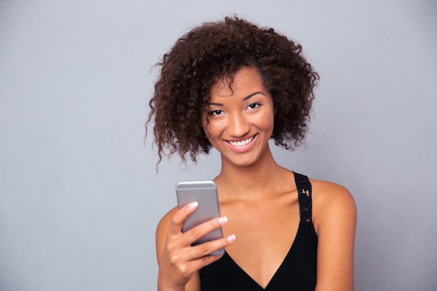 Retrato de una alegre mujer afroamericana con smartphone sobre pared gris y mirando al frente