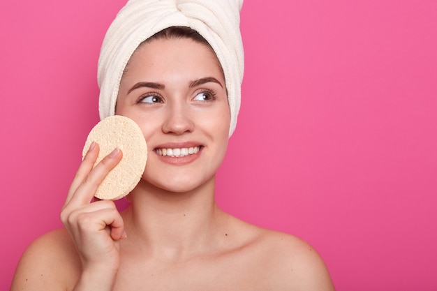 Retrato de alegre mujer caucásica, envuelta en una toalla blanca, limpia la piel con una esponja cosmética