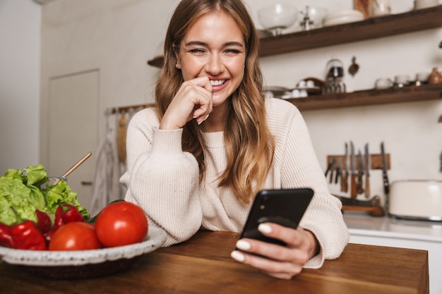 Retrato de alegre mujer caucásica vistiendo ropa casual con teléfono móvil mientras se cocina la cena en la acogedora cocina