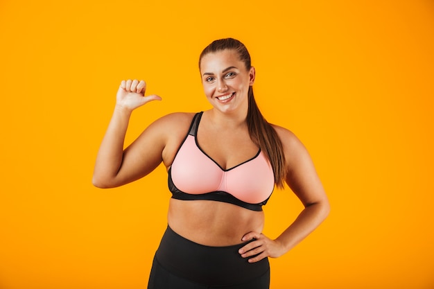 Retrato de una alegre mujer de fitness con sobrepeso vistiendo ropa deportiva que se encuentran aisladas sobre la pared amarilla, apuntando