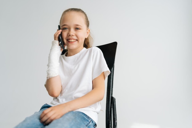 Retrato de una alegre niña rubia con un brazo roto envuelto en una venda de yeso hablando por teléfono inteligente mirando a la cámara sentada en una silla sobre fondo blanco