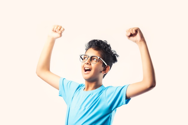 Retrato de alegre niño asiático indio con gafas celebrando el éxito de pie contra la pared blanca