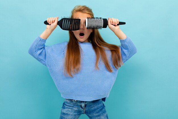 Retrato de una alegre shkolnitsa contenta con cepillos para el cabello en sus manos sobre un fondo azul, cierra los ojos con cepillos para el cabello, tonta